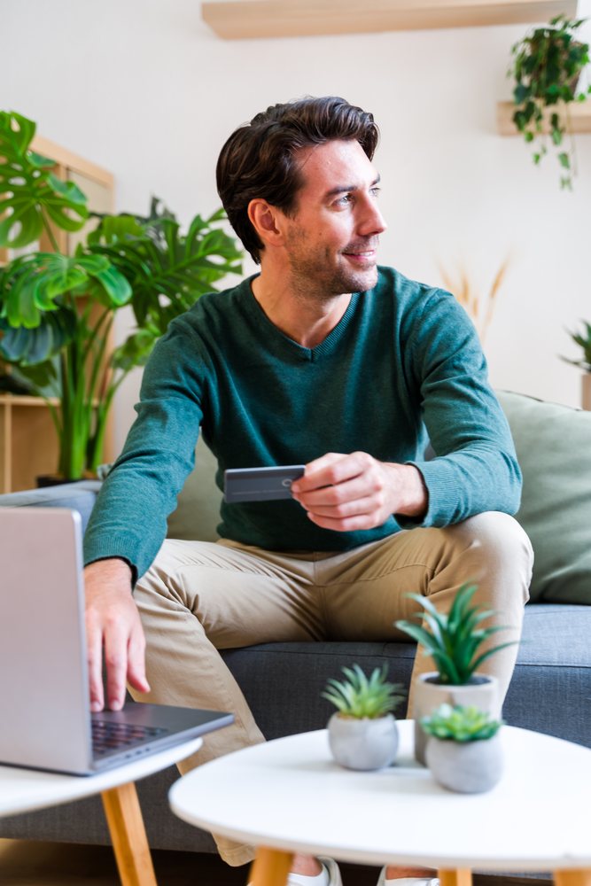 Man using a laptop and holding a credit card while seated in a modern living room with indoor potted plants and a white round table, Spain.