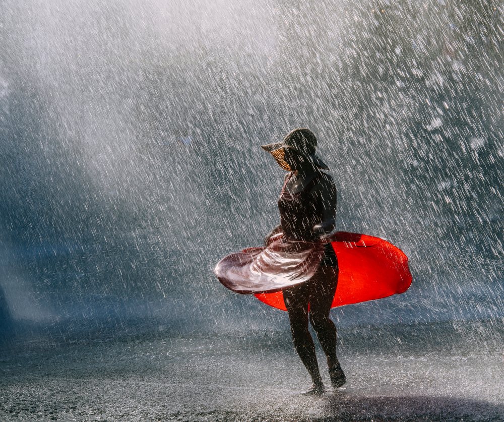 Woman wearing a red dress and sun hat dances in a water spray outdoors, USA.