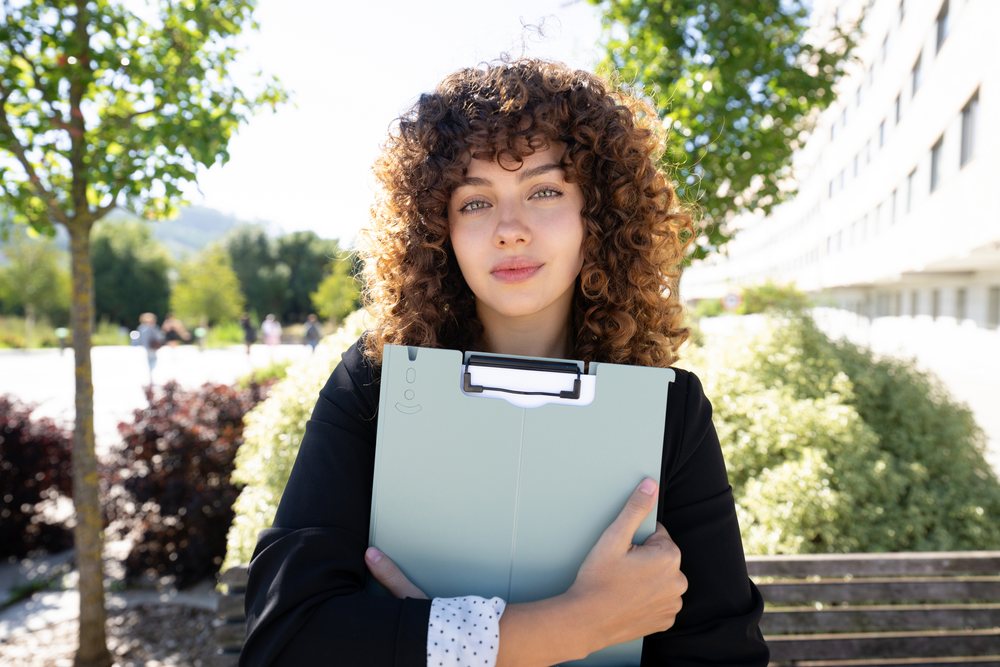 Woman with curly hair holding a clipboard sitting on a park bench amid green trees on a sunny day, Spain.