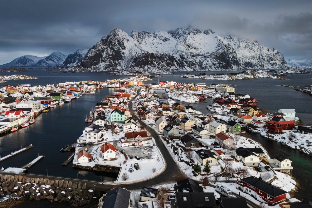 Aerial view of snowy Henningsvaer town with surrounding snow-covered mountains, water channels, and boats docked in harbor, Landscape, Lofoten, Norway.