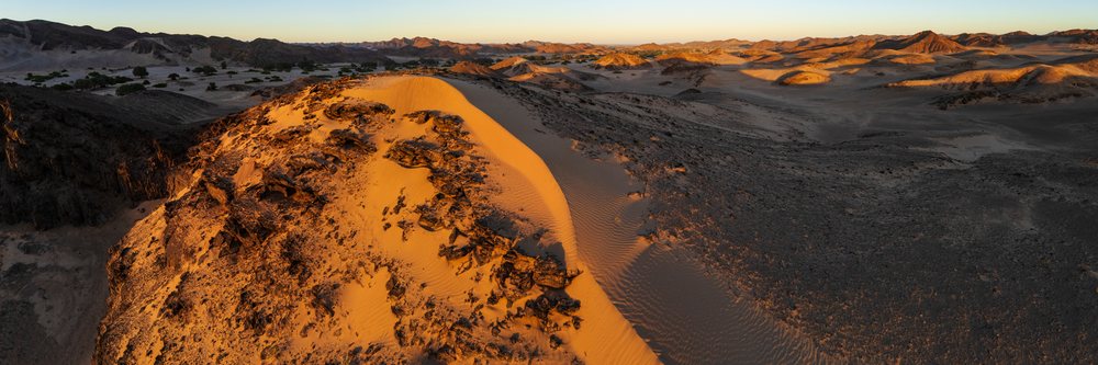 Sunrise illuminating the Hoanib River Valley desert landscape with sand dunes, rock formations, savanna trees, and distant mountains, Kunene Region, Namibia.