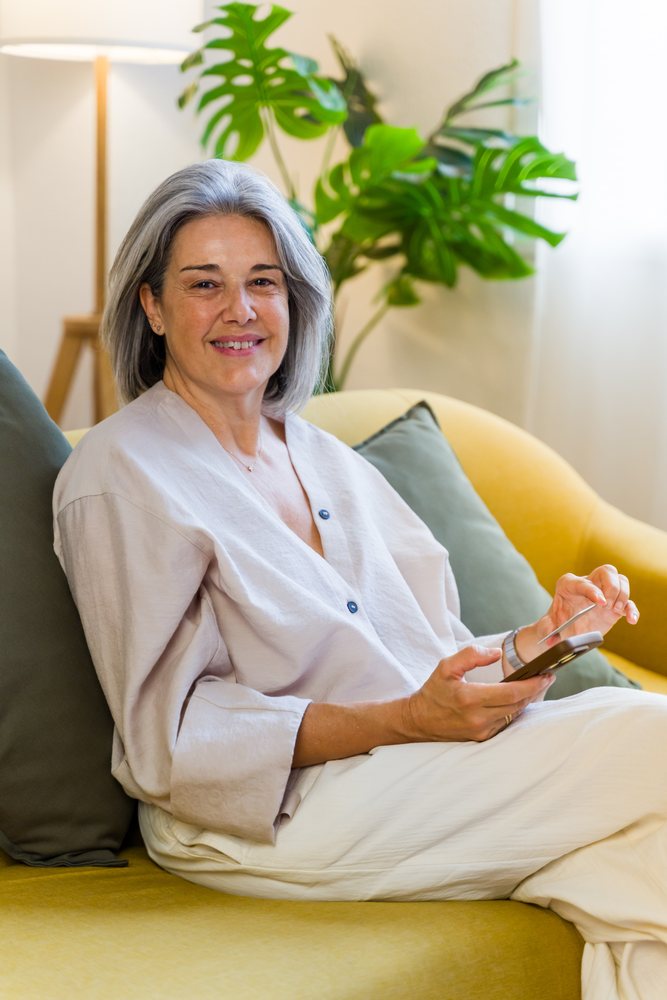 Middle-aged woman with grey hair sits on a yellow couch using a tablet in a living room with indoor plants and natural light, Spain.