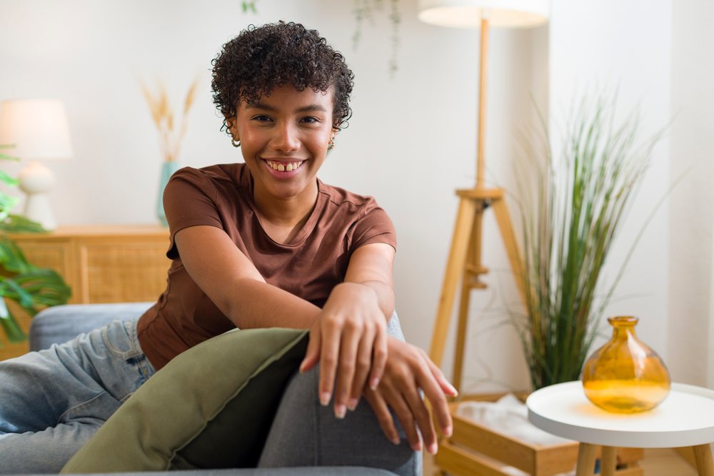 Young woman smiling and relaxing on a couch in a modern indoor living room with wooden furniture and houseplants.