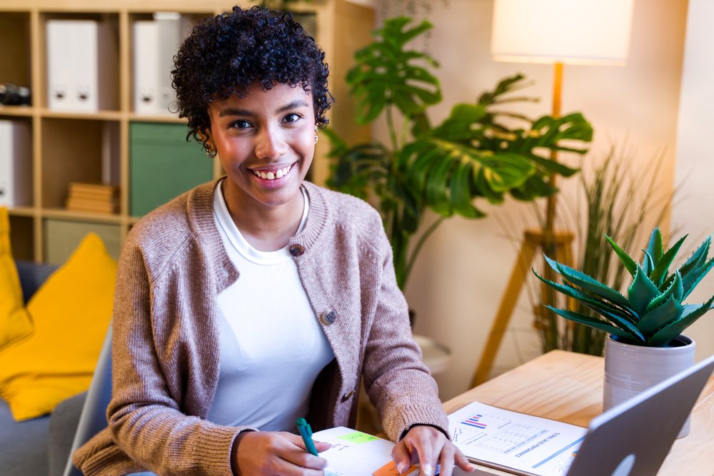 Young woman with curly hair writing in planner at a wooden desk in a bright, modern home office with plants and laptop, Spain.