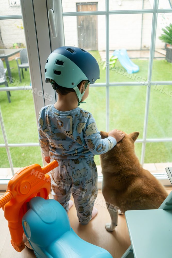 A toddler white boy wearing a helmet stands with his dog inside a home