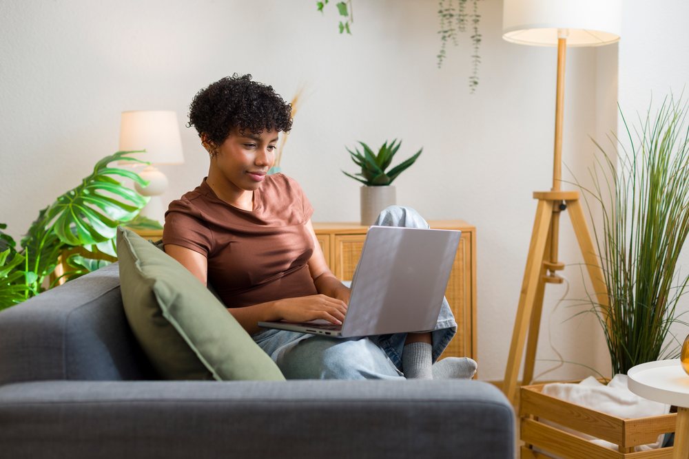 Young woman sits on a sofa using a laptop in a modern living room with plants and a floor lamp.