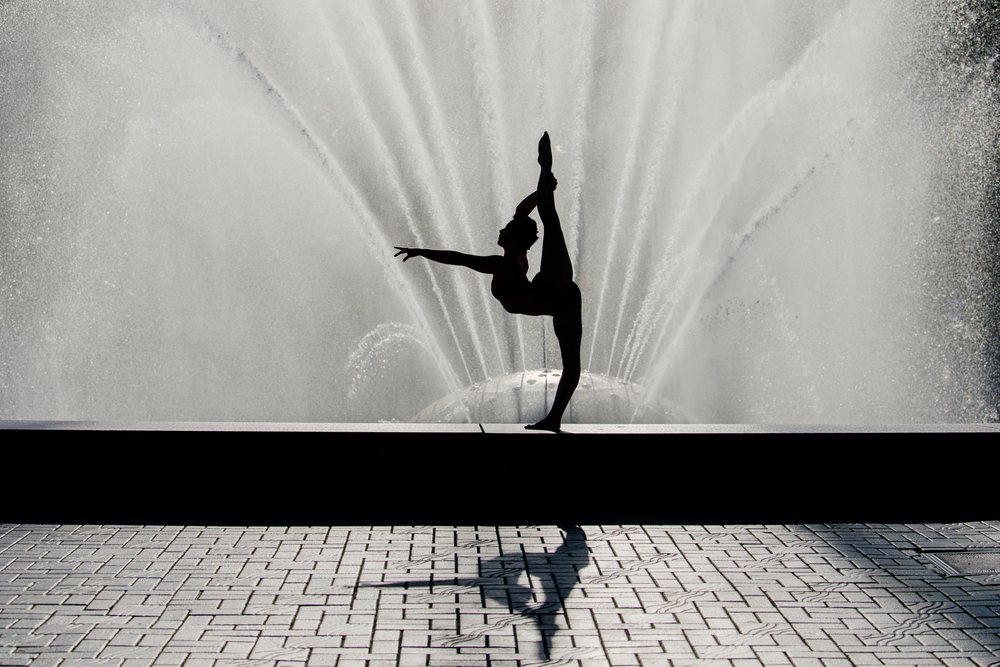 Silhouetted dancer stretches leg high before a water fountain outdoors, USA.