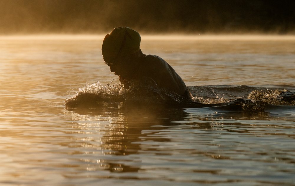 Silhouetted swimmer wearing a swim cap and goggles swimming through misty water at sunrise, USA.