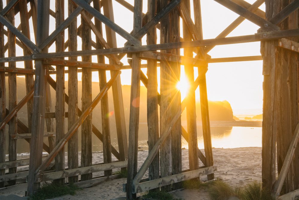 Sunlight passes through wooden beach structure beams with sand and ocean water visible beyond on a northern Oregon coastline, California, USA.