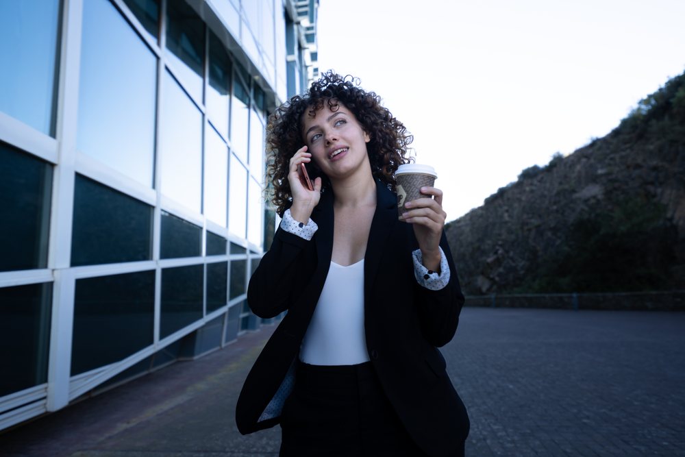 Businesswoman holding a coffee cup and talking on the phone while walking outside a glass building, business trip, Spain, Santiago De Compostela.