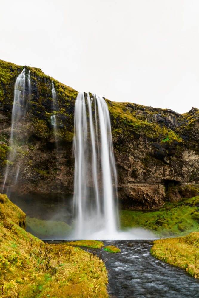 A powerful waterfall cascades over a moss-covered cliff into a flowing river in a lush green landscape, Iceland.