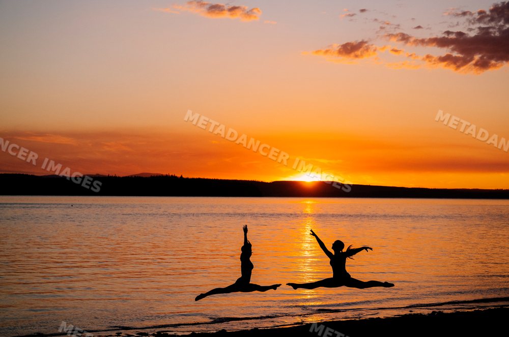 Two white people leap in yoga partner acrobatics by a lake at sunset
