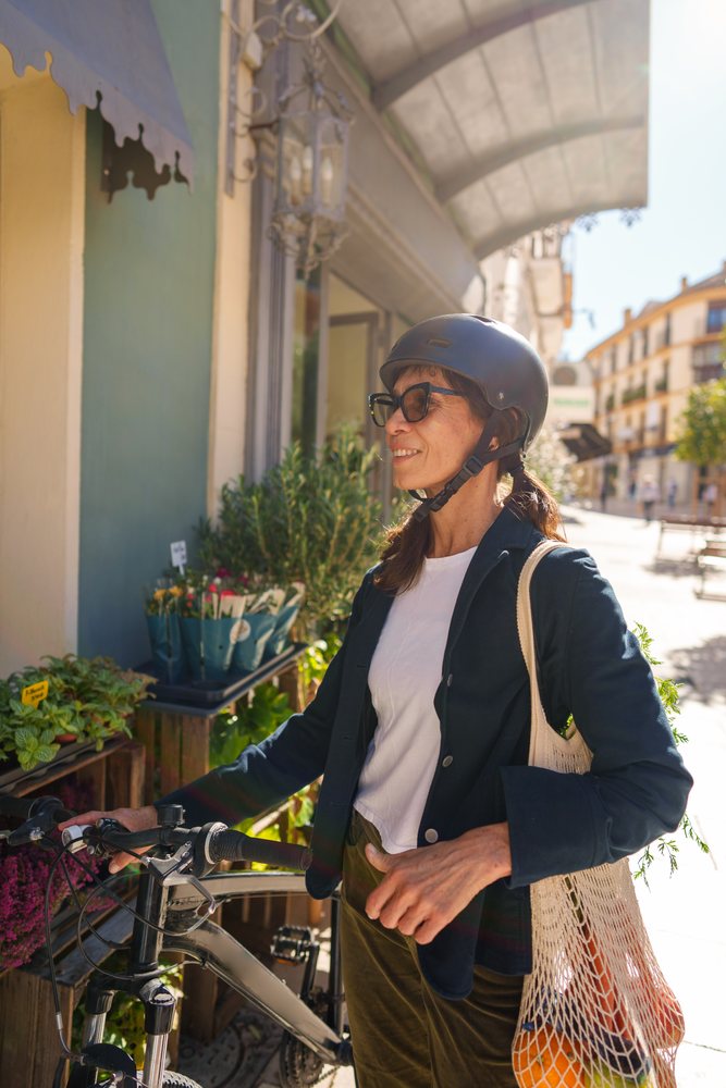 Woman wearing a black helmet and sunglasses holds a bicycle and carries a string shopping bag with oranges at an outdoor market on a sunny day, Málaga.