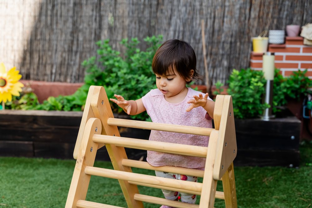 Toddler girl playing on a wooden climber playground toy in a garden with green plants and artificial grass outdoors, kids, Spain.