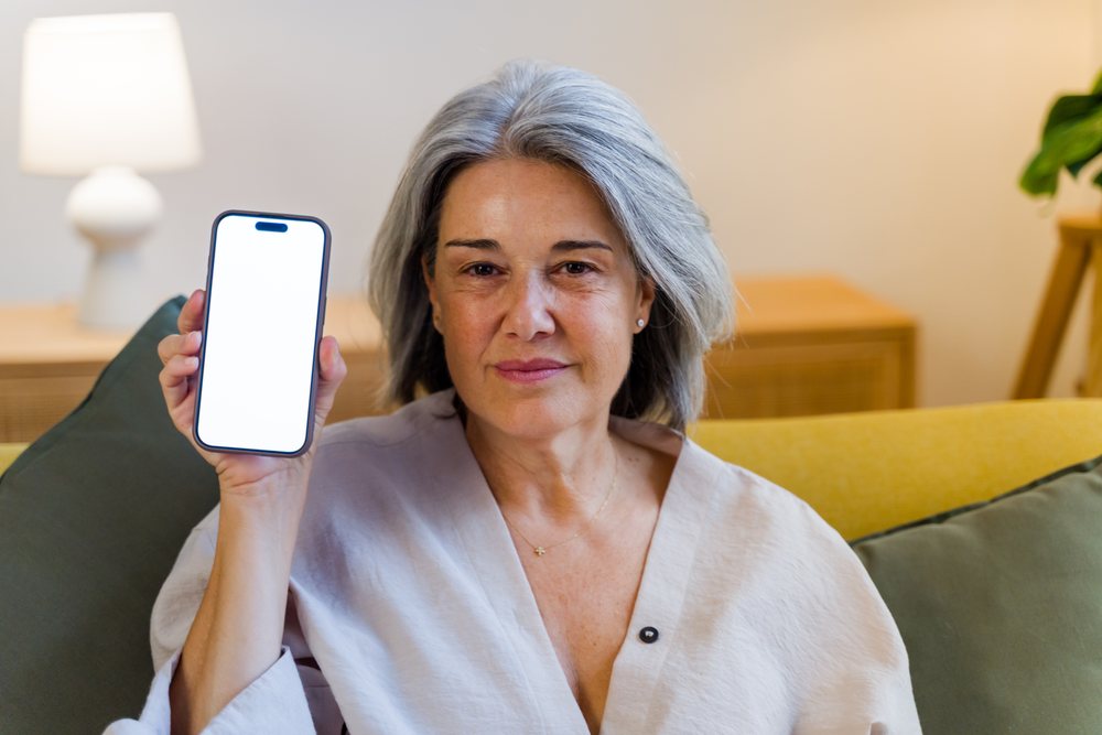 Woman with gray hair wearing light-colored top holding smartphone with blank screen while seated on sofa with lamp and potted plant, Spain.