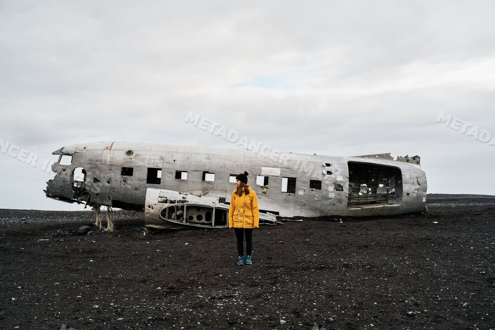 A white woman stands by crashed airplane wreckage on volcanic terrain