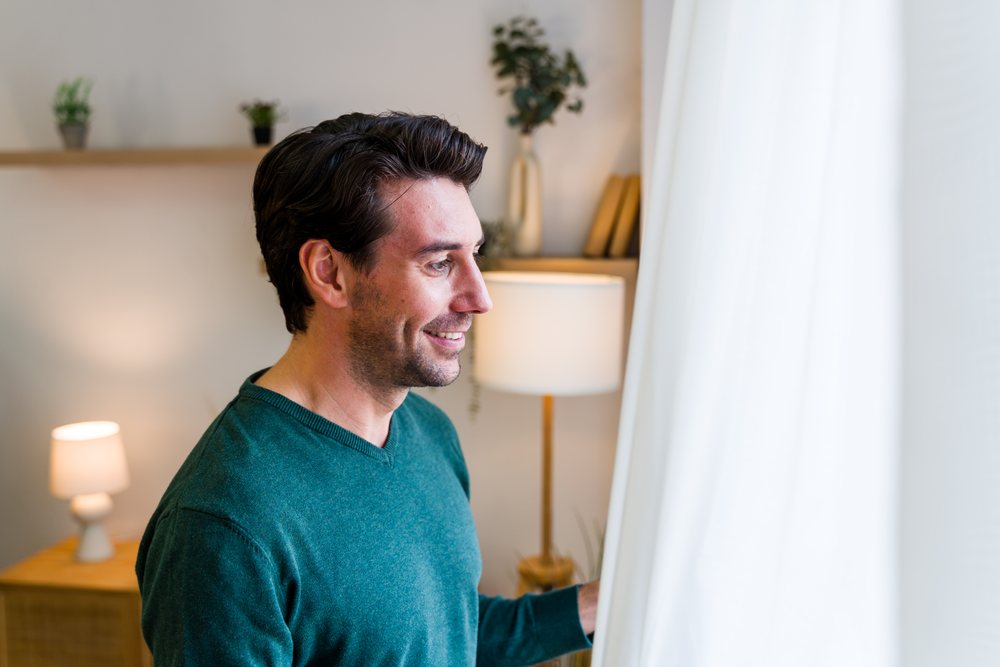 Man wearing a green sweater smiles while looking out the window in a cozy living room, Spain.