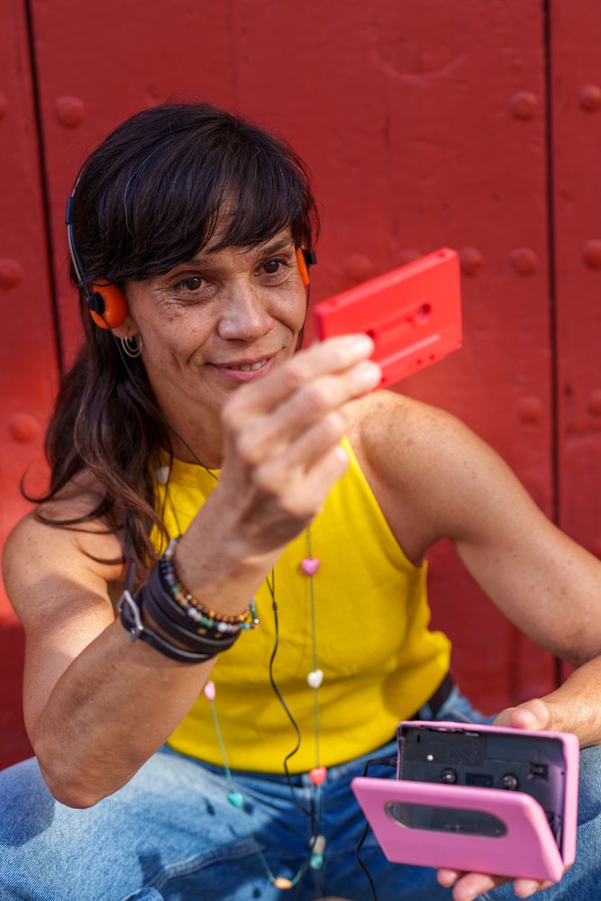 Woman smiling and listening to music on a cassette player with headphones while sitting outdoors by a red wall, retro, vintage, Málaga, Spain.
