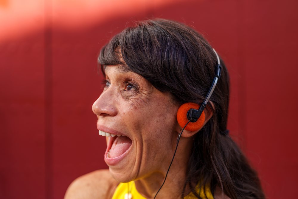 Profile of a woman wearing orange headphones and a yellow sleeveless shirt talking with a surprised expression against a red background, retro, vintage, Málaga, Spain.