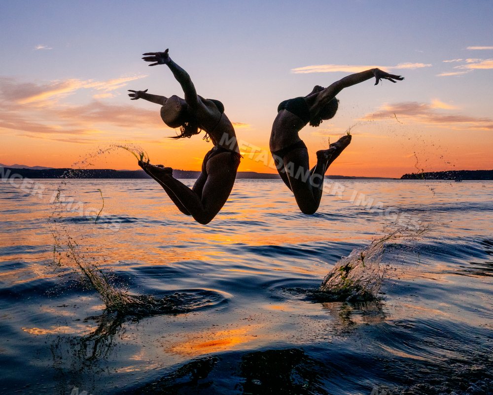 Two women perform partner acrobatics yoga jumps over water at sunset