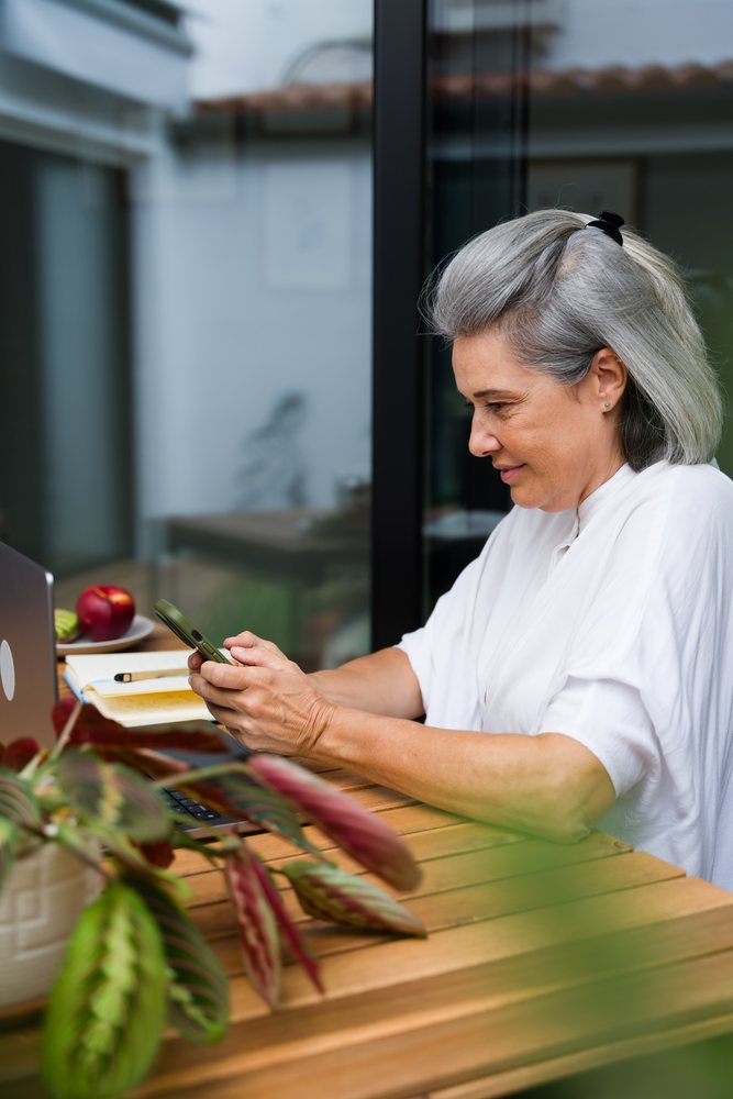 Mature woman with gray hair using a smartphone while seated at a wooden table with a laptop, notebook, and apple in a modern home office, Spain.