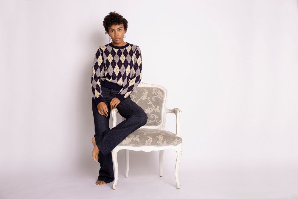 Young woman with curly hair wearing an argyle sweater and dark jeans poses barefoot seated on a decorative chair in a white studio background, fashion, young culture.