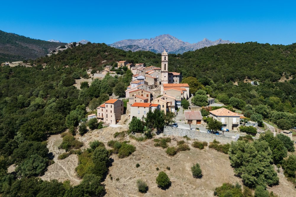 Soveria village on a hilltop, with stone houses, a church tower, and forested mountains under a clear blue sky, Travel Photography, Corsica, France.