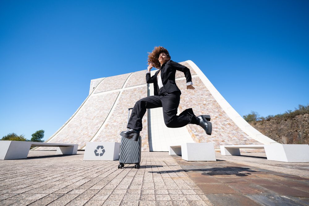 Woman jumping with suitcase in front of modern architecture in an urban plaza, woman, traveling professional, Spain, Antiago De Compostela.