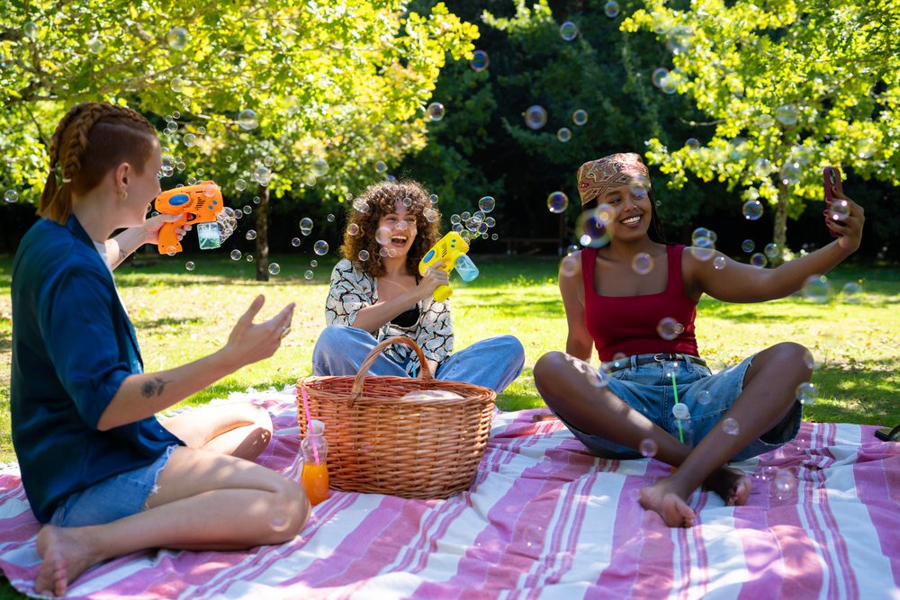 Friends enjoy a picnic in a sunny grassy park playing with bubble guns and taking a selfie, Spain.