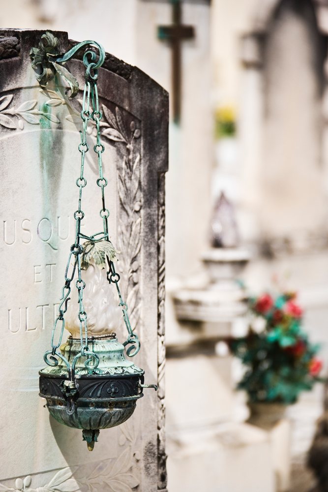 Stone headstone with carved leaf decoration, metal ornamental lamp on chains, and nearby flower decoration in the historic cemetery, Florence, Italy.