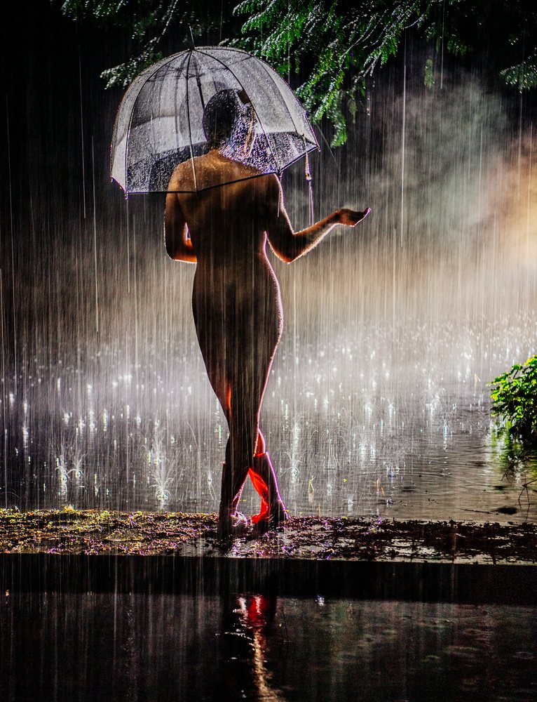 Nude woman wearing rain boots holds a transparent umbrella standing in heavy rain on wet ground with tree reflections at night, cosplay, nudity, freedom, expression, USA.