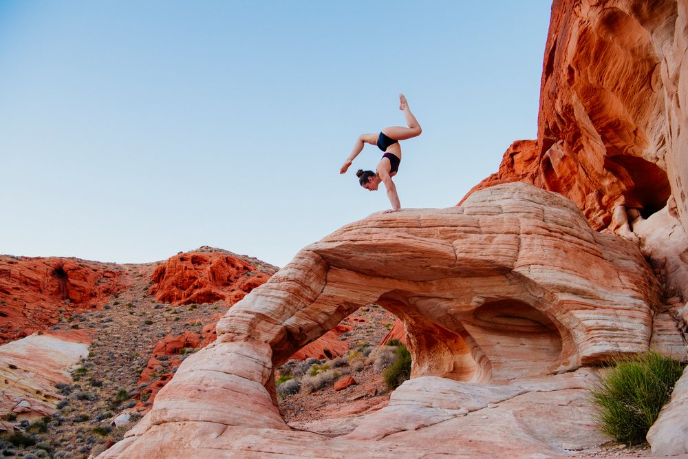 Woman performing a handstand yoga pose on a natural red rock arch in a desert landscape, USA.