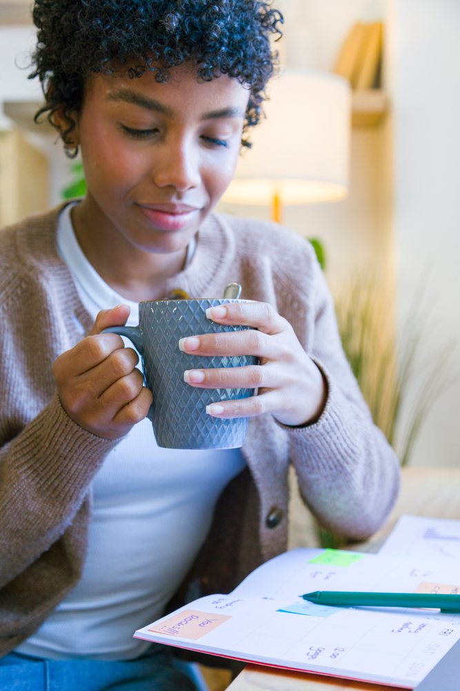 Woman holding a gray ceramic mug seated at a table with a planner and pen in a home office setting, remote work.