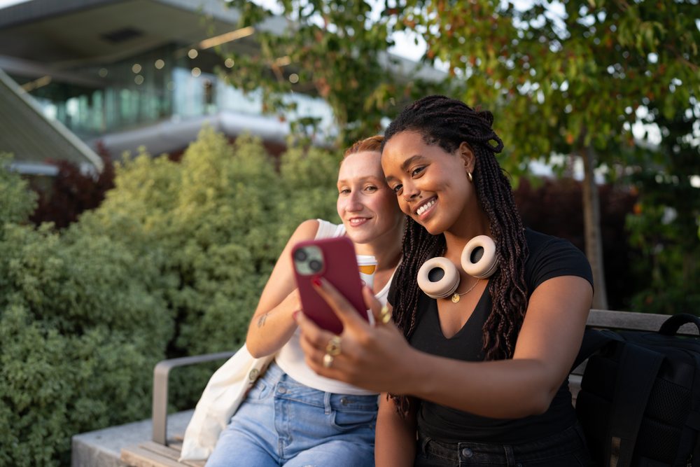 Two women sitting on an outdoor bench taking a selfie with a smartphone near green bushes, urban, Santiago De Compostela, Spain.