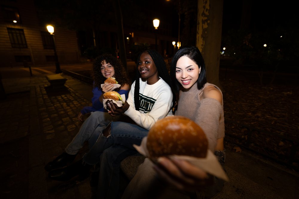 Three women smiling and holding hamburgers while sitting on a bench in a park at night under street lamps, Madrid, Spain.