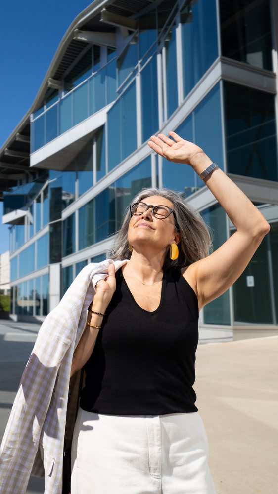 Woman with gray hair wearing glasses and a black sleeveless top holding a beige checkered jacket over her shoulder outdoors in bright sunlight at a modern business district, businesswomen, Spain.