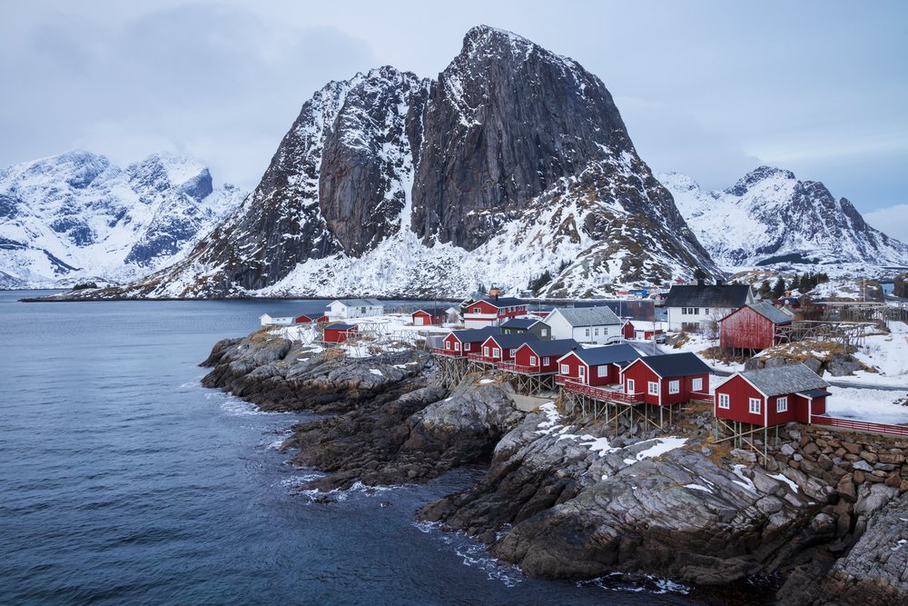 Red houses along a rocky coastline with snow and a large mountain in the background, Landscape, Hamnoy, Lofoten, Norway.