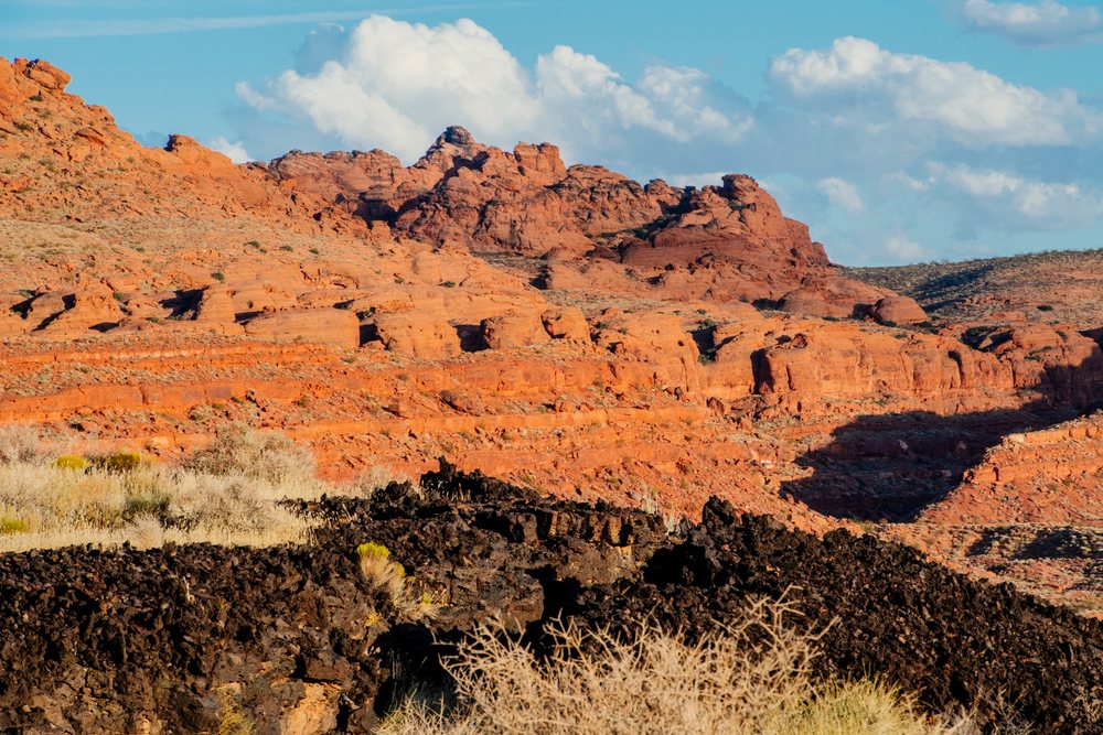 Red rock formations and volcanic rocks rise above desert vegetation under a blue sky with clouds at red Cliffs Desert State Park, Snow Canyon, Utah, USA.