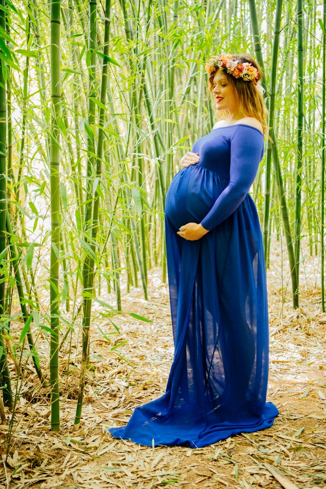 Pregnant woman wearing a blue dress and flower crown stands peacefully among bamboo stalks in an outdoor forest setting with green leaves and dry leaf litter, pregnancy.
