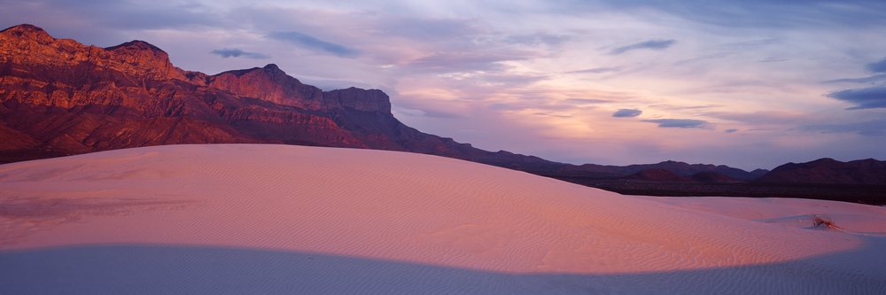 Sand dunes and rugged desert mountains under a colorful sunset sky with clouds.