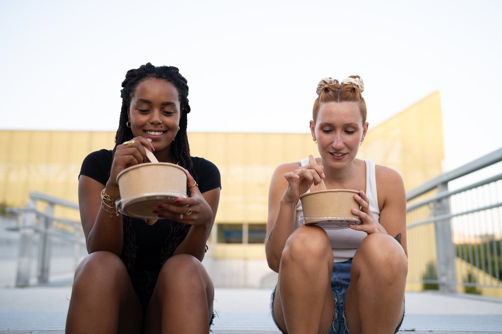 Two young women sit outdoors eating from paper food bowls in an urban setting, Santiago De Compostela, Spain.