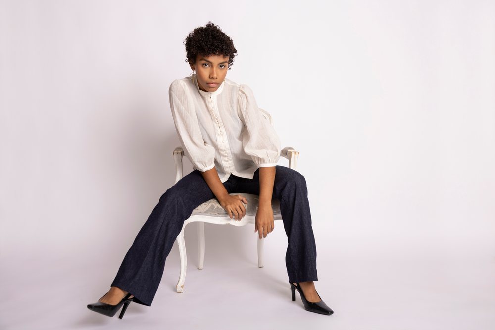 Woman sitting on a chair in a studio wearing a white blouse and dark pants, fashion, young culture.