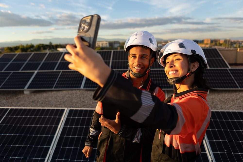 Two workers in white hard hats and orange-black uniforms take a selfie outdoors at a solar panel installation, Spain under a partly cloudy sky, Madrid.
