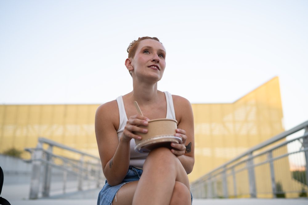 A young woman in casual clothing enjoys eating from a food container while sitting outdoors in an urban setting, Santiago De Compostela, Spain.