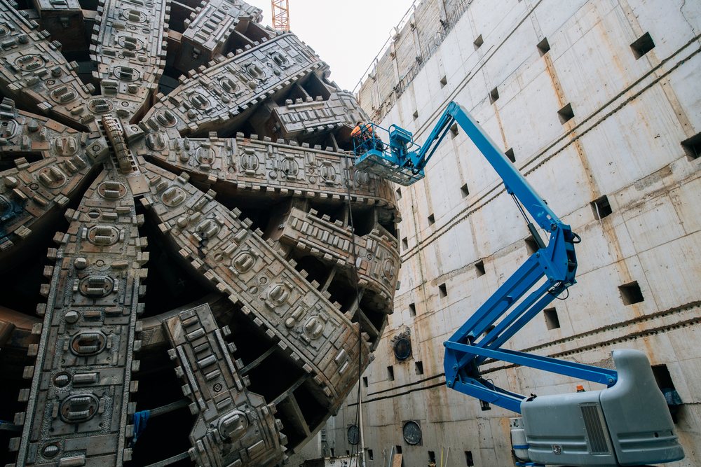 A construction worker inspects a large tunnel boring machine using a blue boom lift inside a concrete structure, Bertha, drilling, USA.