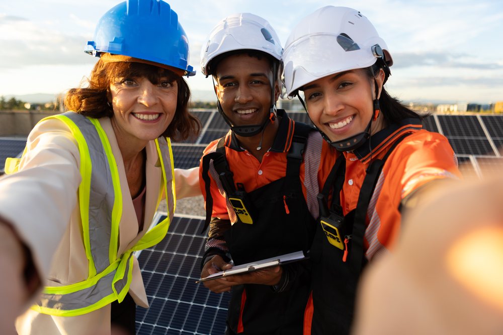 Three workers wearing helmets and safety vests pose for a selfie at a solar panel installation site outdoors in Madrid during daytime, Spain.