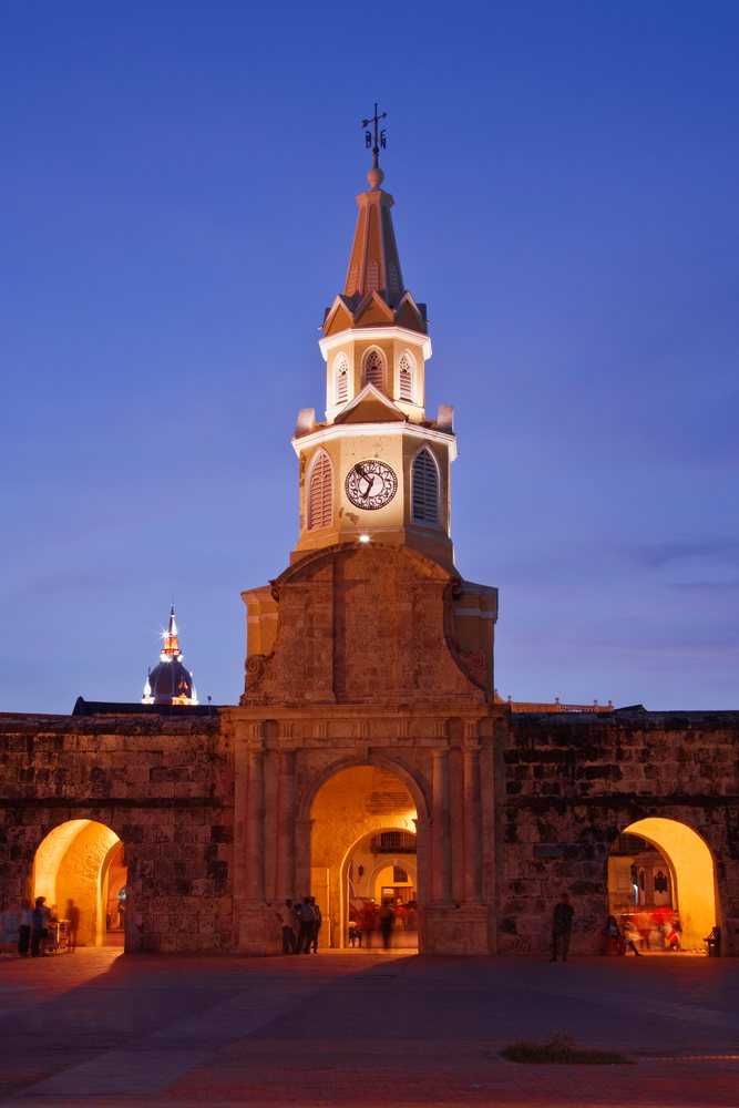 The Puerta del Reloj historic clock tower and stone arch gate are illuminated at night with people nearby under a blue twilight sky, Cartagena, Colombia.