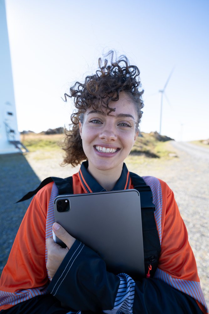 Smiling woman with curly hair wearing a safety vest holds a tablet outdoors at a wind farm with turbines spinning, sustainability, renewable energy, Galicia, Spain.