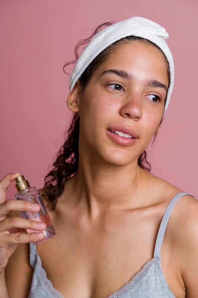 Woman wearing a white headband holds a clear facial spray serum bottle against a pink background during a beauty treatment, Spain.