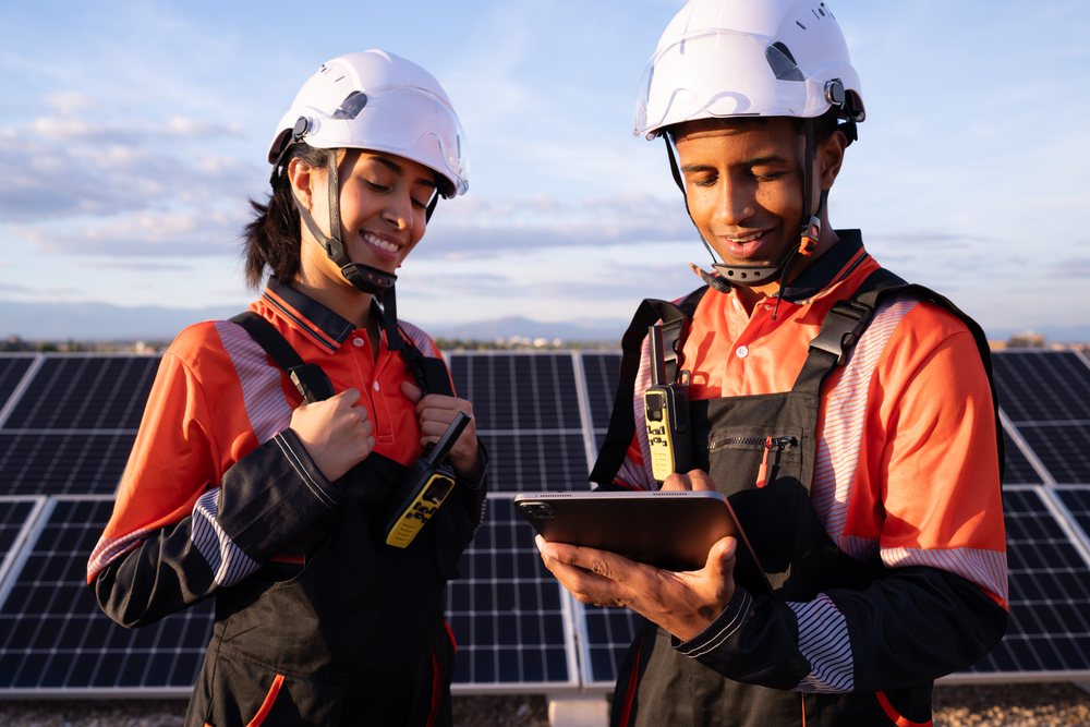 Two technicians wearing safety helmets and protective clothing inspect solar panels outdoors in sunlight, Madrid, Spain.